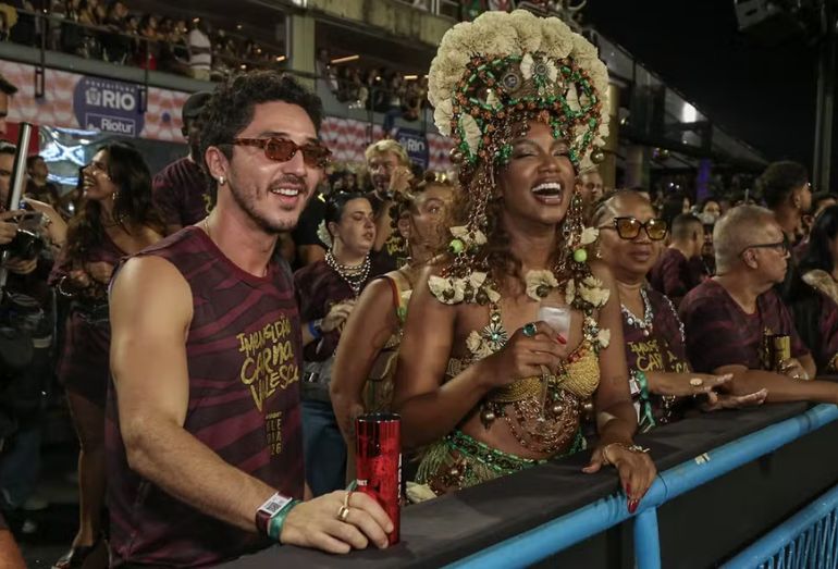 A cantora Iza e o ator João Vitor Silva abraçados e sorrindo em um camarote decorado na Marquês de Sapucaí.