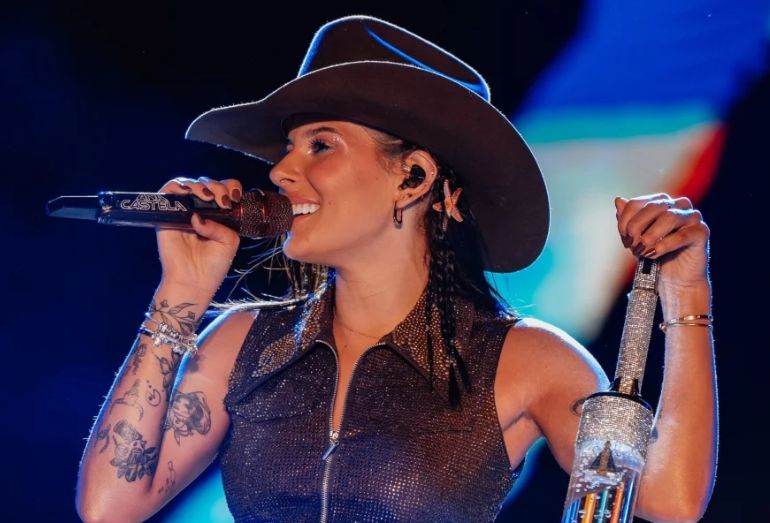 A cantora Ana Castela, usando chapéu e jaqueta de franjas, olhando para o horizonte com um semblante pensativo.