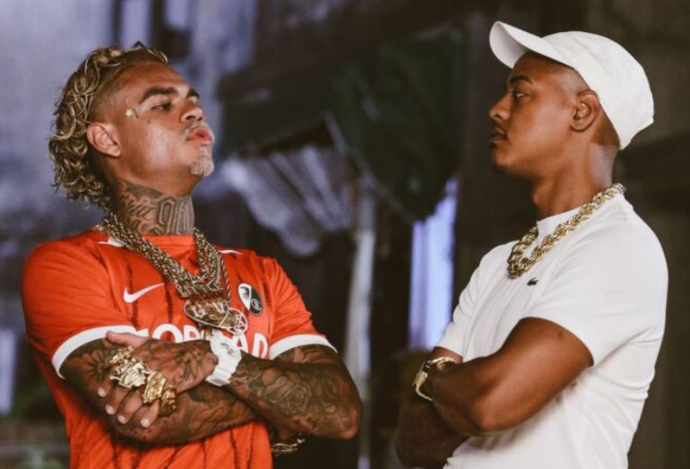 MC Cabelinho e TZ da Coronel posando juntos com estilo urbano e roupas de grife, em um cenário que mistura elementos de luxo e estética de periferia.