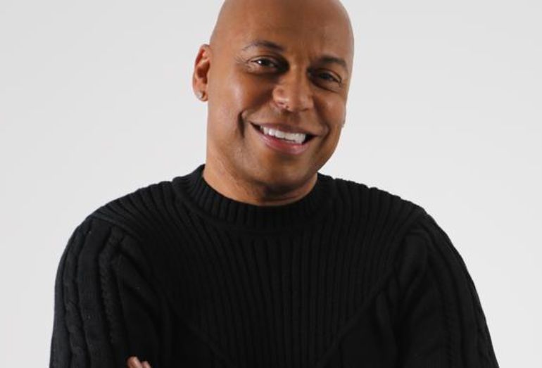 Close-up do compositor Délcio Luiz sorrindo e de braços cruzados, vestindo uma blusa de lã preta, contra um fundo branco.