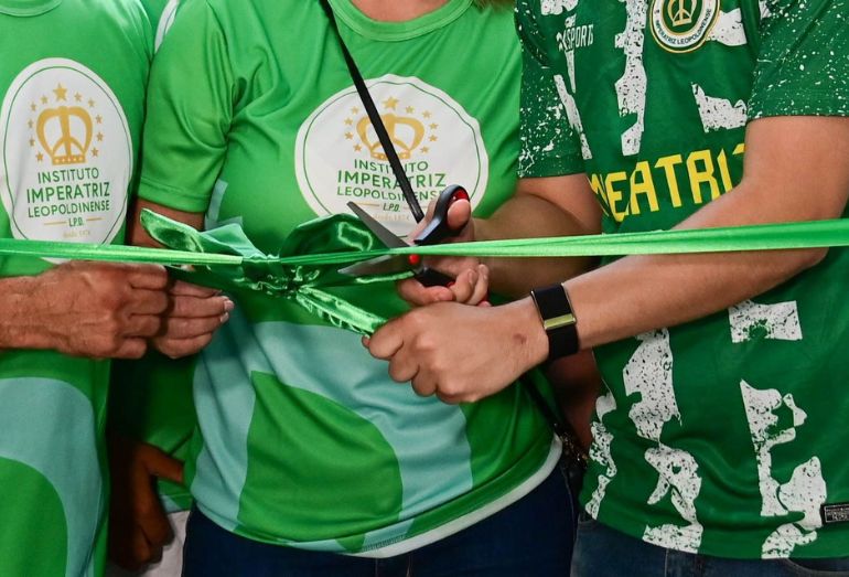 Close-up das mãos de João Drumond e Catia Drumond utilizando uma tesoura para cortar a fita inaugural verde, simbolizando a abertura da sede própria do Instituto Imperatriz Leopoldinense. Eles vestem camisas verdes com a logomarca do instituto.