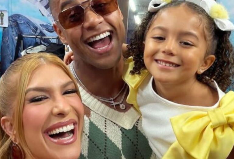 Selfie sorridente da família. Lore Improta aparece em primeiro plano à esquerda, Léo Santana ao centro usando óculos escuros e a pequena Liz à direita com um grande laço amarelo no cabelo. Todos estão rindo e demonstrando felicidade.