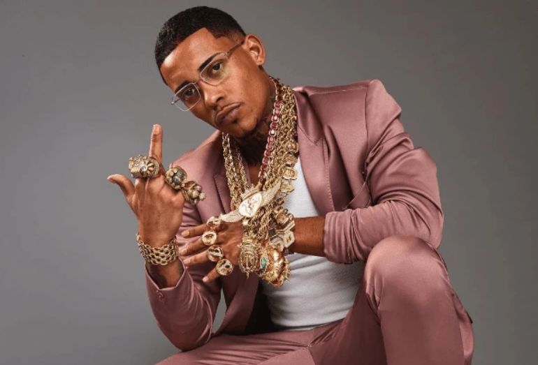 Close de corpo inteiro do cantor MC Poze do Rodo, agachado e sorridente, vestindo um terno rosa metálico sobre uma camiseta branca. Ele está coberto por joias ostensivas, incluindo múltiplas correntes de ouro grosso e pingentes com diamantes, além de anéis e pulseiras de ouro em ambas as mãos.