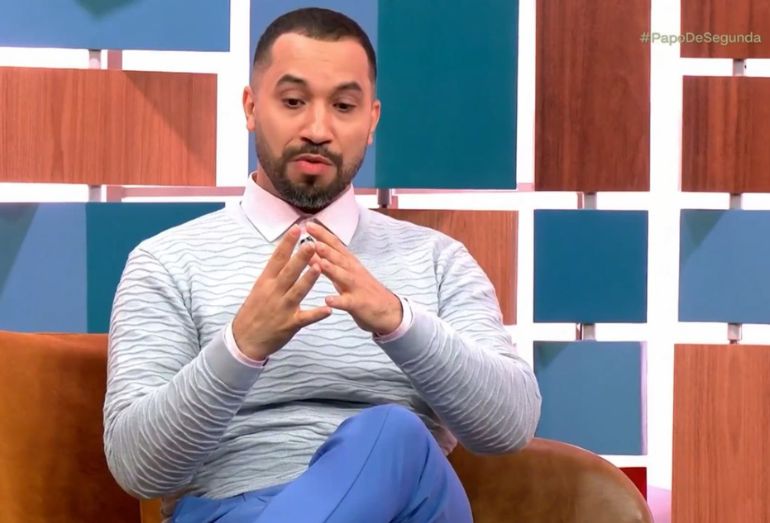 Gil do Vigor sentado no sofá do programa Papo de Segunda, falando de forma expressiva e emocionada.