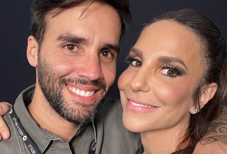 Uma foto de arquivo, em plano médio e close, de Ivete Sangalo e Daniel Cady abraçados e sorrindo para a câmera. Eles estão com as bochechas coladas. Ivete Sangalo está à direita, com os cabelos castanhos presos e maquiagem destacada com delineador escuro e cílios postiços, usando um brinco de brilhante pequeno. Daniel Cady está à esquerda, com barba curta e veste uma camisa de botões verde-oliva sobre uma camiseta preta, usando um crachá de evento pendurado no pescoço. O fundo é escuro e liso.