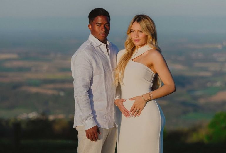Uma fotografia ao ar livre, em plano médio, do jogador Endrick e da influenciadora Gabriely Miranda em uma paisagem de horizonte aberto. Endrick, um jovem negro à esquerda, veste uma camisa social branca de linho e calças claras, olhando seriamente para a câmera. Gabriely, uma mulher branca de cabelos loiros ondulados à direita, veste um vestido longo off-white justo e de um ombro só. Ela está de perfil, olhando para o horizonte, com as mãos delicadamente posicionadas sobre o ventre, sinalizando a gravidez. O fundo apresenta uma vista panorâmica de campos e vegetação sob a luz suave do entardecer.