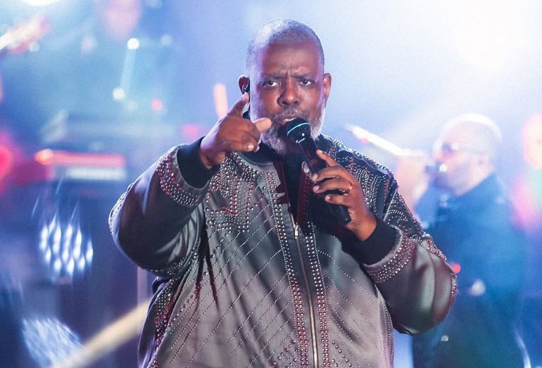 Uma imagem em plano médio de Péricles cantando e apontando para a plateia durante sua apresentação no festival The Town. Veste uma jaqueta bomber marrom-avermelhada customizada com detalhes brilhantes e colares de miçangas vermelhas cobrindo o peito e ombros. Ele segura um microfone na mão esquerda e aponta com o dedo indicador direito. O fundo é composto por luzes de palco dramáticas em tons de azul e branco, criando um efeito de halo e feixes de luz no ar, com outros músicos levemente visíveis e desfocados ao fundo.