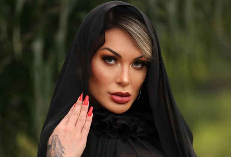 Retrato em close da influenciadora Andressa Urach usando um véu de tecido preto transparente sobre a cabeça e uma gola alta de babados pretos. Ela apresenta uma maquiagem social marcante, unhas longas pintadas de vermelho e uma tatuagem no pulso, mantendo uma expressão séria enquanto encara a câmera em um ambiente externo com fundo verde desfocado.