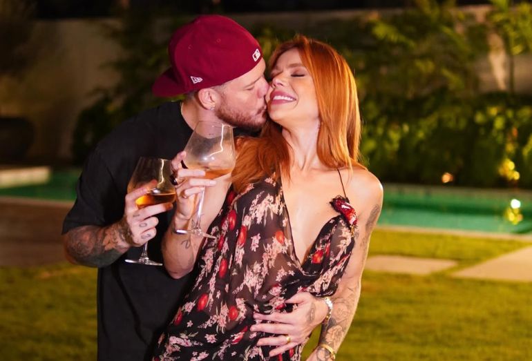 O cantor Ferrugem abraçado com sua noiva Thaís Vasconcellos durante uma festa de aniversário, ela sorri emocionada.