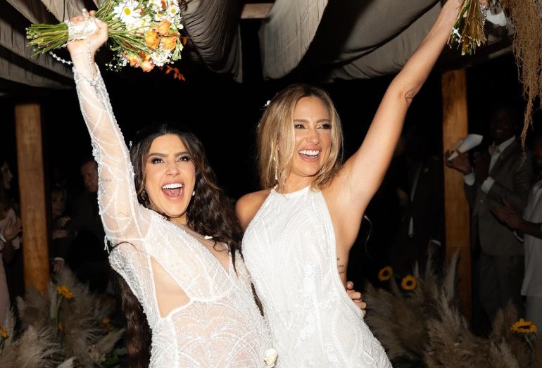 Foto das noivas Lauana Prado e Tati Dias de branco, sorrindo durante o casamento em Ubatuba.