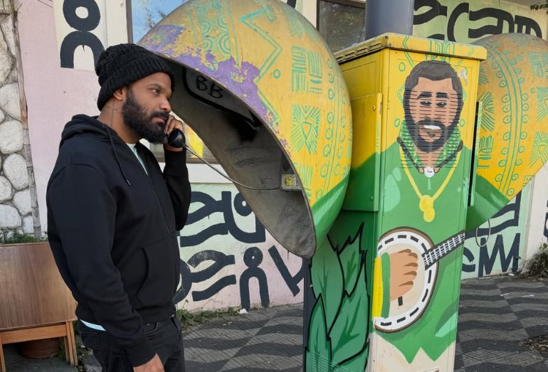 Foto do cantor Arlindinho encostado em um telefone público antigo grafitado com o rosto e o banjo do cantor Arlindo Cruz, olhando para a imagem com expressão de saudade.
