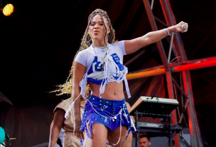 A cantora Marvvila em performance no palco. Ela usa um conjunto azul de top e minissaia com detalhes em jeans e correntes, sobre uma camiseta branca curta. Ela segura o microfone com o braço esquerdo estendido e faz uma expressão confiante enquanto se apresenta.